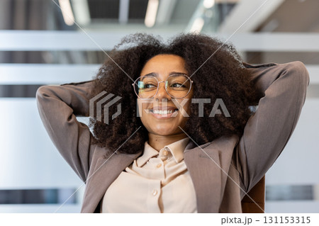 Close-up photo of a young African-American woman in a suit and glasses sitting in an office chair at a desk, hands behind her head and smiling contentedly. Close-up photo of a young African-American woman in a suit and glasses sitting in an office chair at a desk, hands behind her head and smiling contentedly. 131153315