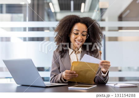 A smiling young African-American businesswoman is sitting at the desk in the office and opens an envelope with a received letter. 131154288
