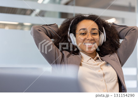Close-up portrait of a satisfied young African-American woman sitting at an office desk in a suit and wearing headphones, hands behind her head and resting, looking and smiling at the camera. Close-up portrait of a satisfied young African-American woman sitting at an office desk in a suit and wearing headphones, hands behind her head and resting, looking and smiling at the camera. 131154290