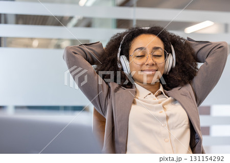 Young smiling African American woman sitting in office wearing headphones and resting, eyes closed and hands behind head. Young smiling African American woman sitting in office wearing headphones and resting, eyes closed and hands behind head. 131154292
