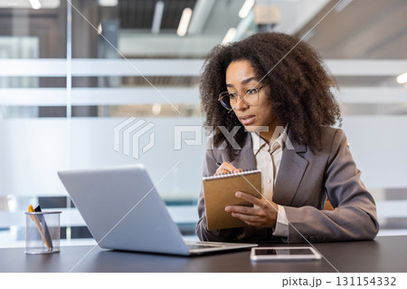 Concentrated young african american woman in business suit sitting in office at desk in front of laptop and taking notes. 131154332