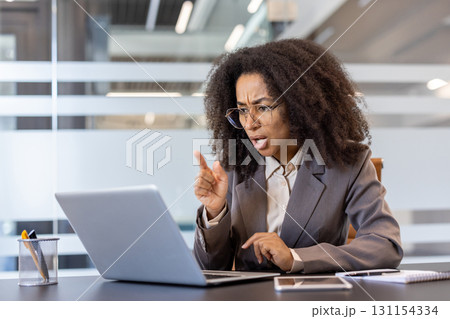 Angry young african american business woman sitting at desk in office and talking angrily on video call on laptop camera while pointing finger at screen. 131154334