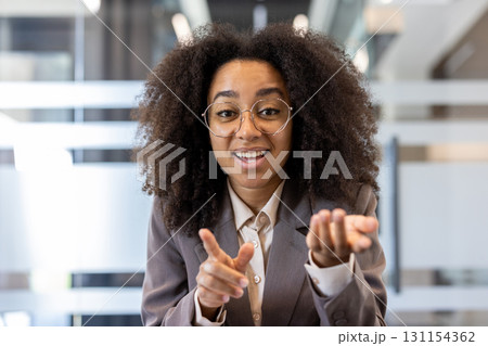 Close-up portrait of a young African-American woman in glasses and a suit sitting in front of the camera in an office , at a desk and talking to the camera while gesturing with her hands. Close-up portrait of a young African-American woman in glasses and a suit sitting in front of the camera in an office , at a desk and talking to the camera while gesturing with her hands. 131154362