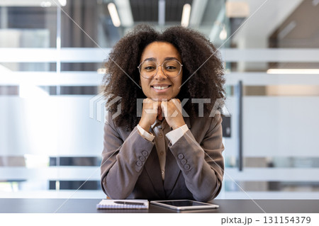 Close-up photo of a smiling African-American female office worker sitting at her desk in a suit, hands under her chin and looking at the camera. Close-up photo of a smiling African-American female office worker sitting at her desk in a suit, hands under her chin and looking at the camera. 131154379