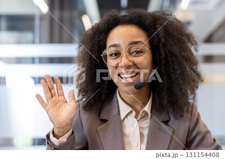 Close-up portrait of a young African-American woman in a suit and glasses wearing a headset sitting in an office in front of the camera, talking and shaking hands. Close-up portrait of a young African-American woman in a suit and glasses wearing a headset sitting in an office in front of the camera, talking and shaking hands. 131154408