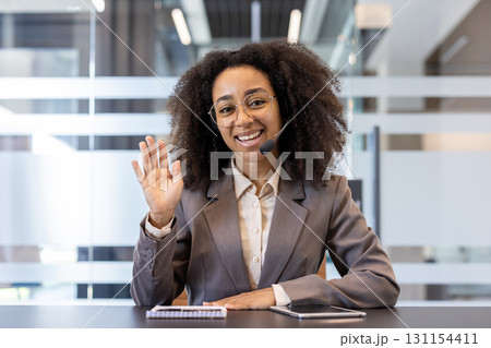 Smiling young African American woman wearing headset and suit sitting in office at desk, talking and greeting to camera. 131154411
