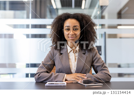 Portrait of a young African American woman in a suit and headset sitting in the office at a desk, looking and smiling at the camera. Portrait of a young African American woman in a suit and headset sitting in the office at a desk, looking and smiling at the camera. 131154414