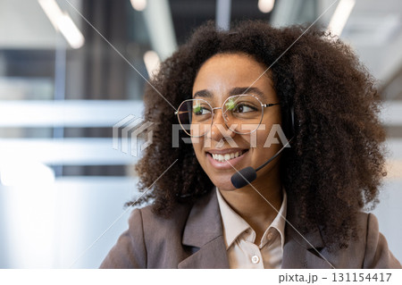 African American young woman in a headset, suit and glasses is in an office building, working and studying, smiling and looking to the side. Close-up photo. African American young woman in a headset, suit and glasses is in an office building, working and studying, smiling and looking to the side. Close-up photo. 131154417