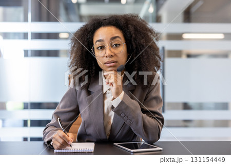 Portrait of a young African American businesswoman sitting in the office in front of a camera in a headset, making notes in a notebook and holding an online meeting. Portrait of a young African American businesswoman sitting in the office in front of a camera in a headset, making notes in a notebook and holding an online meeting. 131154449