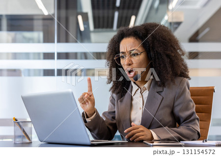 Angry and serious young African American woman in headset and suit talking emotionally on video call on laptop gesturing with hand. Angry and serious young African American woman in headset and suit talking emotionally on video call on laptop gesturing with hand. 131154572