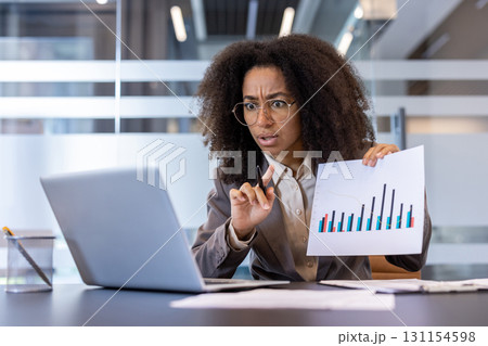 Worried young African American woman sitting in office at laptop, holding online meeting and angrily showing documents and graphs on screen. Worried young African American woman sitting in office at laptop, holding online meeting and angrily showing documents and graphs on screen. 131154598