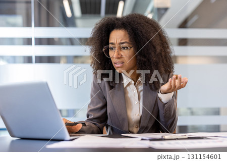 Frustrated young african american business woman sitting in office at work desk with documents and looking at laptop screen in frustration with arms outstretched. Frustrated young african american business woman sitting in office at work desk with documents and looking at laptop screen in frustration with arms outstretched. 131154601