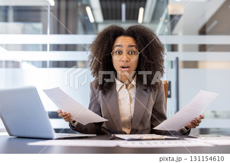 Portrait of disappointed and upset young African-American woman sitting in the office at the desk, holding documents and papers, throwing up her hands in frustration. 131154610