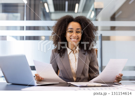 Portrait of a smiling young African American woman in a suit sitting at a desk in the office, holding documents and papers in her hands and looking at the camera. 131154614