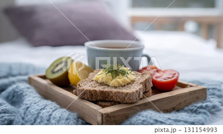 Healthy breakfast tray with whole grain toast, kiwi, lemon, tomato, and herbal tea on cozy bed 131154993