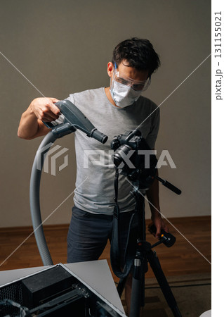 Vertical portrait of professional videographer wearing protective mask and glasses sanitizing camera and computer equipment. Concept of computer hardware, repairing, upgrade and technology. 131155021