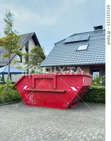Large red dumpster filled with debris placed outside a modern home on a cloudy day in a suburban neighborhood 131155520