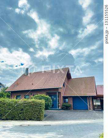 Modern suburban house with a red tiled roof and blue garage under a cloudy sky in a quiet neighborhood Modern suburban house with a red tiled roof and blue garage under a cloudy sky in a quiet neighborhood 131155526