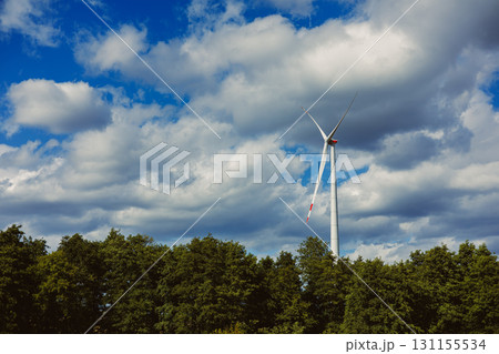 Large wind turbine standing tall among green trees under a bright blue sky with fluffy clouds Large wind turbine standing tall among green trees under a bright blue sky with fluffy clouds 131155534