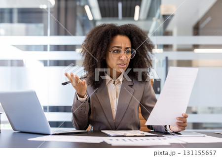 Upset young African-American woman working in the office, sitting at the desk, looking disappointedly at the documents in her hand. 131155657