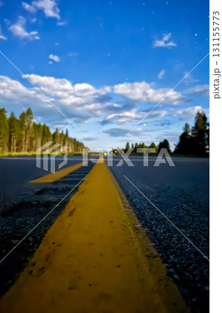 Stretching yellow lines lead down a wide open road under a bright blue sky filled with fluffy clouds during golden hour before dusk 131155773