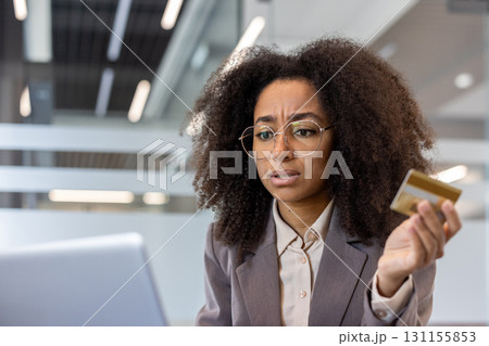 Upset young African American woman sitting at desk in office, holding credit card and looking frustrated at laptop screen. 131155853