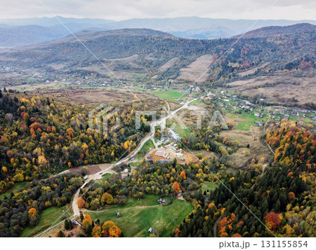 Aerial view of tranquil valley in autumn. Vibrant fall foliage and small village nestled among rolling hills. Winding roads connect scattered houses. Carpathian mountains, Ukraine. Aerial view of tranquil valley in autumn. Vibrant fall foliage and small village nestled among rolling hills. Winding roads connect scattered houses. Carpathian mountains, Ukraine. 131155854