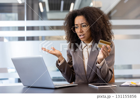 Credit card problems. African american young upset woman sitting in the office at the desk and looking disappointedly at the laptop and holding a plastic bank card in her hand. 131155856