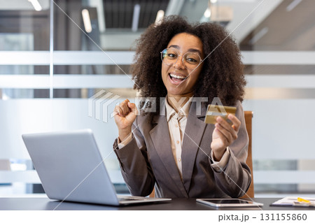 Portrait of a young African American business woman in glasses and a suit sitting at the workplace in the office and happy holding a credit card in her hands. Portrait of a young African American business woman in glasses and a suit sitting at the workplace in the office and happy holding a credit card in her hands. 131155860