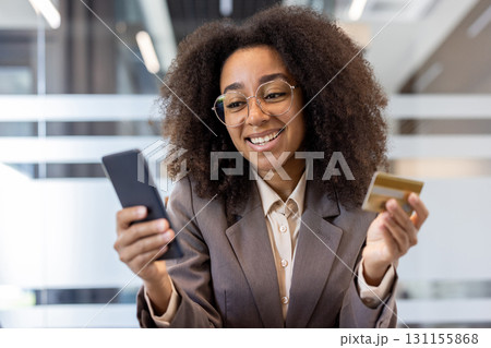 Close-up photo of a young African-American woman in a suit and glasses sitting in an office, holding a credit card and looking at a mobile phone screen. 131155868