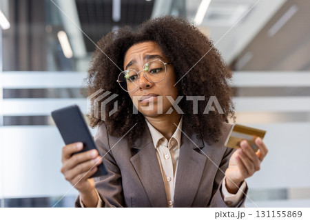 Close-up photo of a sad young African-American woman sitting at a desk in the office, holding a credit card and looking frustrated at the phone screen. Close-up photo of a sad young African-American woman sitting at a desk in the office, holding a credit card and looking frustrated at the phone screen. 131155869