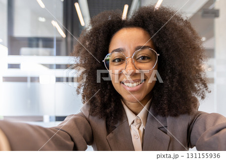 Close-up portrait of a young smiling and beautiful African American woman in a business suit and glasses holding a camera, and talking on a video call. Close-up portrait of a young smiling and beautiful African American woman in a business suit and glasses holding a camera, and talking on a video call. 131155936