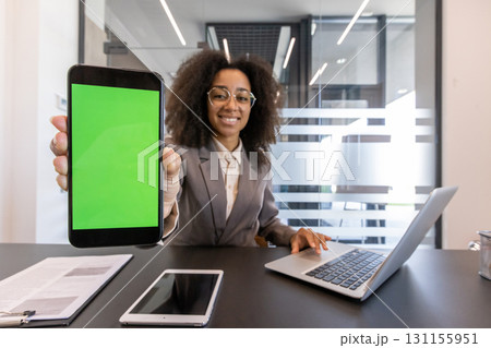 Portrait of a smiling young African-American businesswoman sitting in the office at a desk with a laptop, and showing a green screen mock-up phone to the camera. 131155951