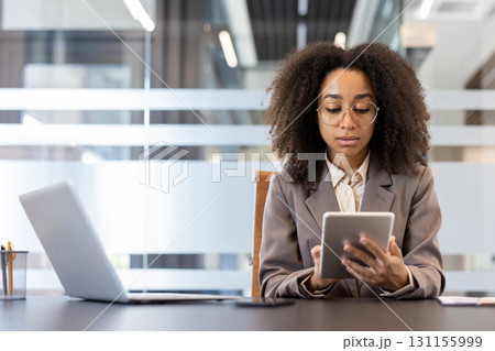 Young African American businesswoman using tablet at workplace inside office. Focused professional with tablet and laptop working efficiency, showcasing modern work environment 131155999