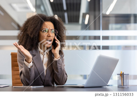 Angry young African American business woman sitting in the office at the desk and emotionally talking on the mobile phone. 131156033
