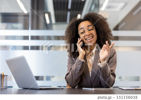 Smiling and successful young African American business woman sitting in the office at the desk and talking on the phone while gesturing with her hands. Smiling and successful young African American business woman sitting in the office at the desk and talking on the phone while gesturing with her hands. 131156038