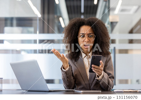 Portrait of an angry young African American woman sitting at a desk in the office, holding the phone and worriedly spreading her hands at the camera. 131156070