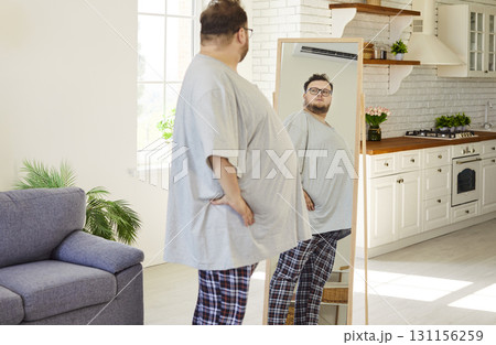 Overweight man examining belly in home mirror, planning diet and lifestyle changes 131156259