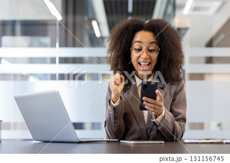 Young African American businesswoman in formal attire celebrating success at workplace inside office holding phone with laptop in front showing excitement and joy 131156745