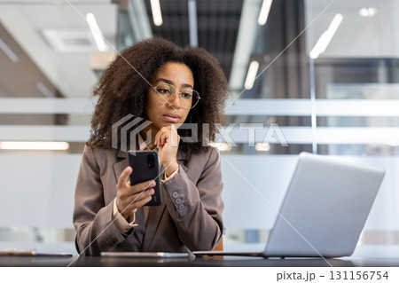 Serious and thoughtful young African American business woman sitting in the office at the desk, holding the phone in her hand and looking at the laptop screen. 131156754