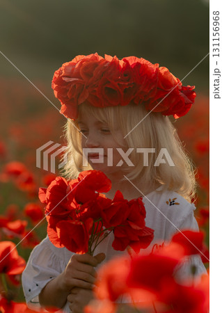 A young girl wearing a red flower crown is standing in a field of red poppies. She is holding a bouquet of red flowers in her hand 131156968