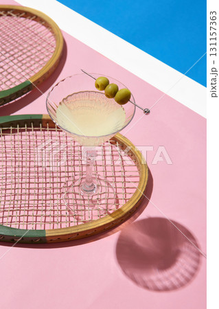 Martini glass with olives on vintage tennis racket over pink surface Martini glass with olives on vintage tennis racket over pink surface 131157363