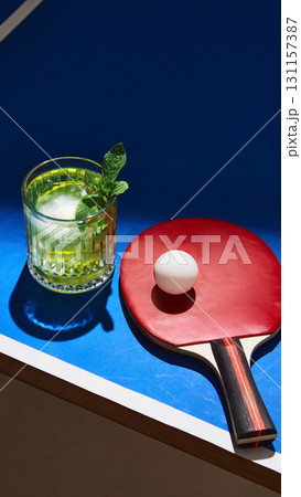Green cocktail with ice and mint beside table tennis paddle and ball on blue background Green cocktail with ice and mint beside table tennis paddle and ball on blue background 131157387