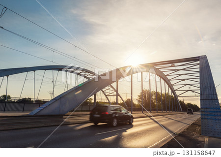Scenic dynamic view of Magdeburg steel arch Jerusalem bridge cars crossing under warm sunrise sunlight. Urban composition highlights architectural structure traffic movement modern city atmosphere Scenic dynamic view of Magdeburg steel arch Jerusalem bridge cars crossing under warm sunrise sunlight. Urban composition highlights architectural structure traffic movement modern city atmosphere 131158647