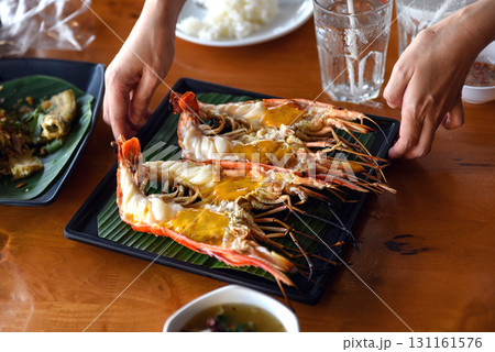 A close-up photo of a hand supporting a plate of large grilled shrimp that had been cut in half, showing oozing fat. Placed on a green banana leaf A close-up photo of a hand supporting a plate of large grilled shrimp that had been cut in half, showing oozing fat. Placed on a green banana leaf 131161576