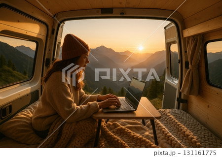 Woman working on laptop from cozy camper van with mountain sunset view in the background during evening hours 131161775