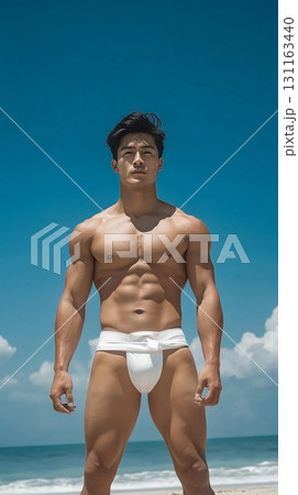 Fit Asian man in swim shorts standing on a tropical beach with a clear blue sky 131163440