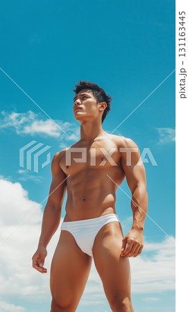 Fit Asian man in swim shorts standing on a tropical beach with a clear blue sky 131163445