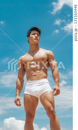 Fit Asian man in swim shorts standing on a tropical beach with a clear blue sky 131163446