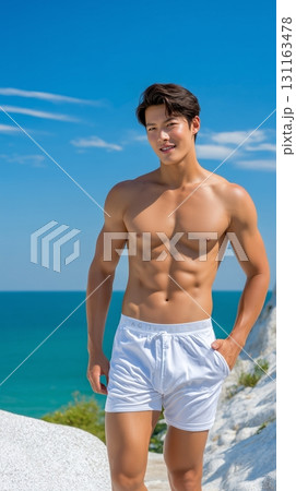 Fit Asian man in swim shorts standing on a tropical beach with a clear blue sky 131163478